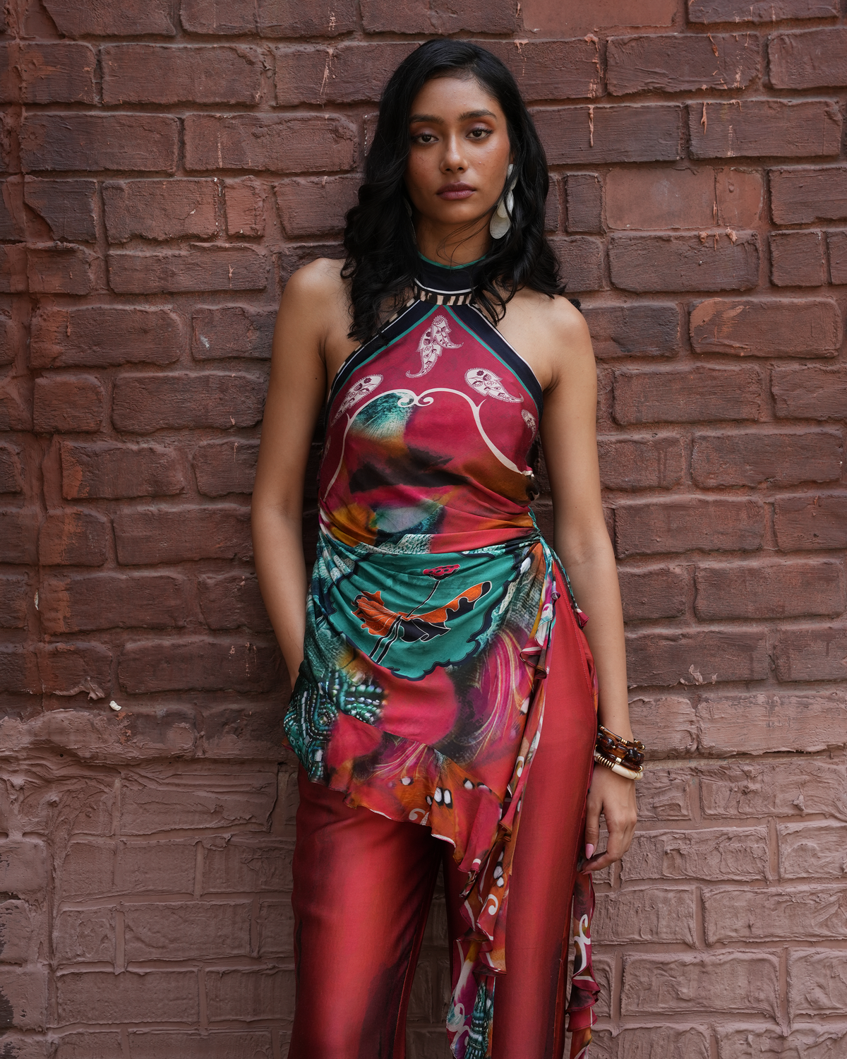 A woman stands against a red-brown brick wall wearing a sleeveless halter-neck dress with a rich red, teal, and orange floral print, paired with matching red pants.