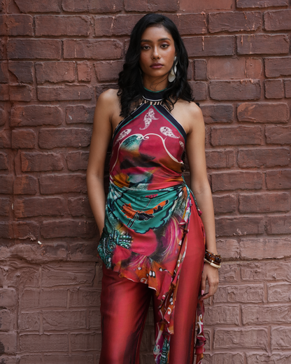 A woman stands against a red-brown brick wall wearing a sleeveless halter-neck dress with a rich red, teal, and orange floral print, paired with matching red pants.