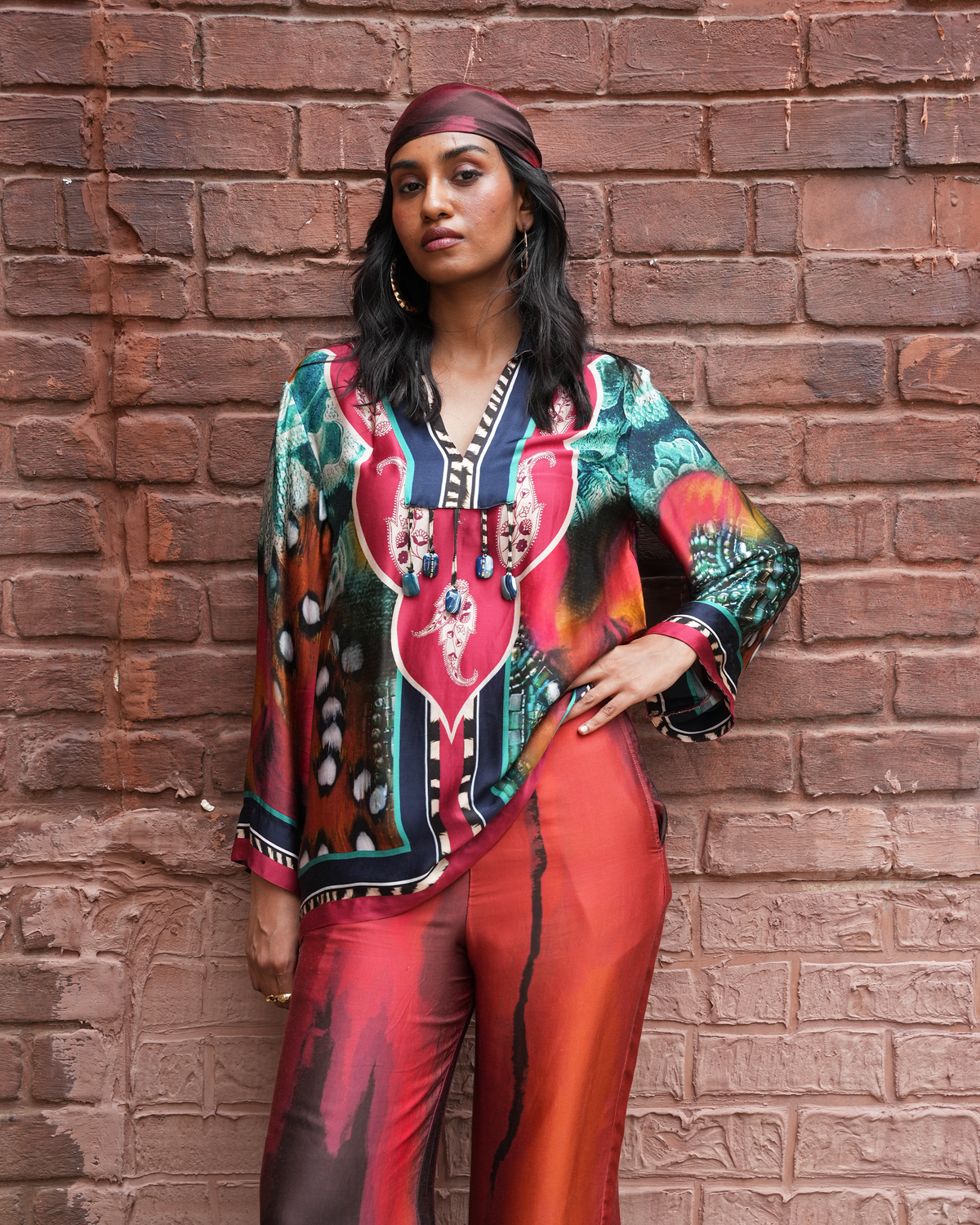 A woman posing against a red-brown brick wall wearing a vibrant, multicolored printed kaftan top with a matching headscarf and bright red-orange satin trousers.