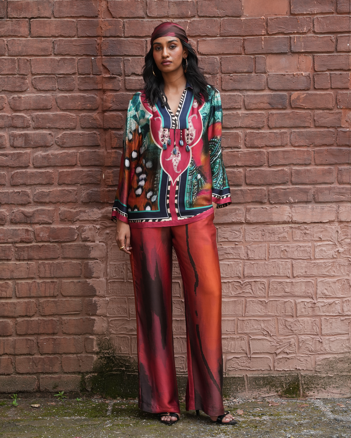A woman stands against a red-brown brick wall wearing a matching two-piece outfit with a vibrant bohemian-style print. The set features a long-sleeve top and wide-leg pants in shades of red, orange, green, and black, accessorized with a dark red headband.