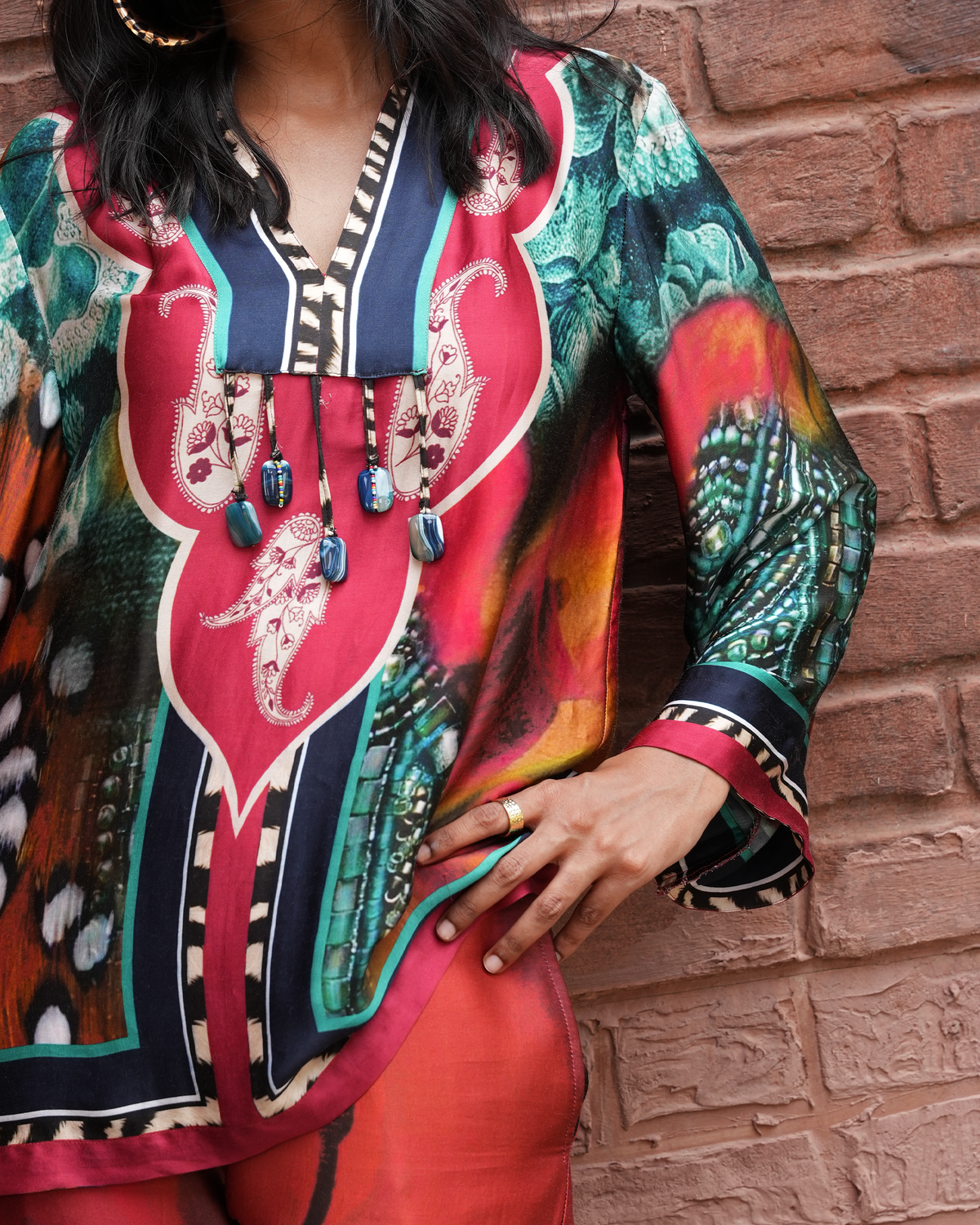 A woman wearing a vibrant, multicolored printed tunic top with a V-neck and decorative beaded necklace, standing against a rustic red brick wall.