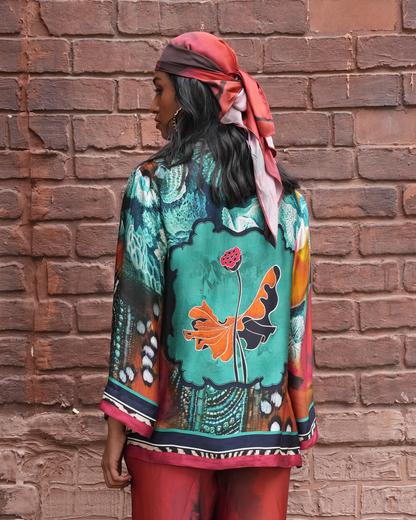 A woman stands with her back to the camera wearing a vibrant, multicolored patterned kaftan-style top and matching red pants, set against a rustic red brick wall.