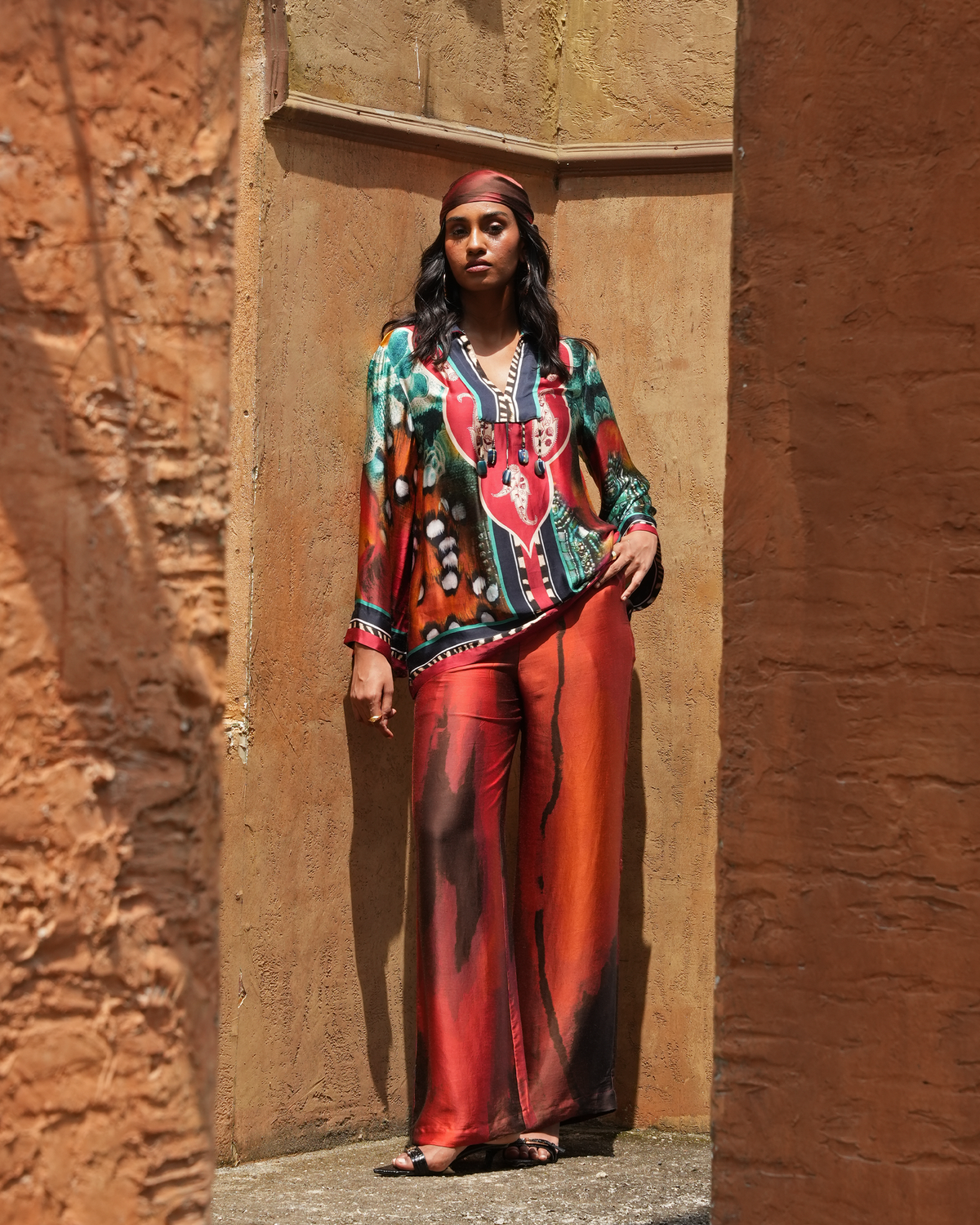 A woman stands in a rustic stone alcove wearing a vibrant, multicolored printed two-piece outfit with a red and green patterned long-sleeve top and matching red wide-leg pants.