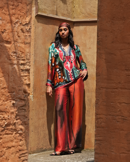 A woman stands in a rustic stone alcove wearing a vibrant, multicolored printed two-piece outfit with a red and green patterned long-sleeve top and matching red wide-leg pants.