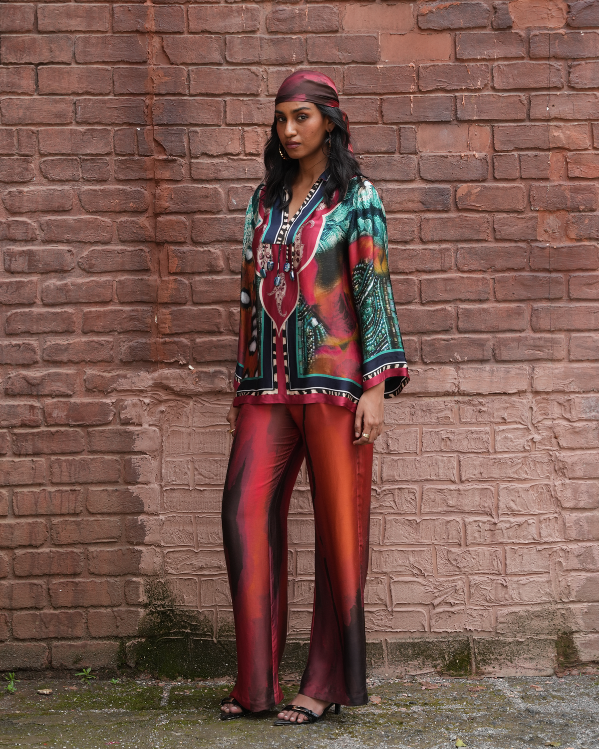 A woman stands against a red-brown brick wall wearing a matching printed outfit with a red headwrap, a multicolor patterned blouse, and red-orange satin-like wide-leg pants.