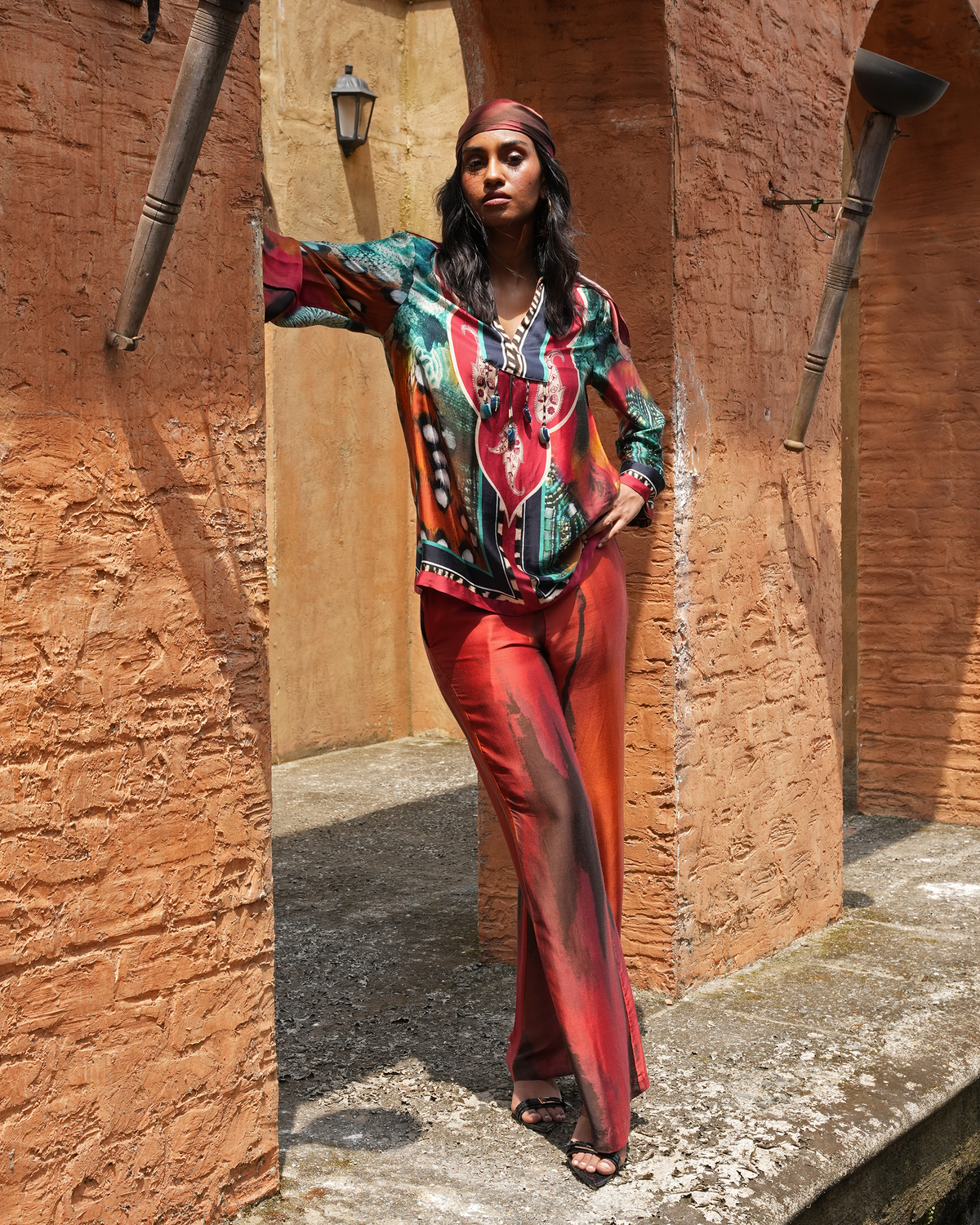 A woman models a vibrant, multicolored bohemian-style printed top and matching red-orange wide-leg pants in an outdoor setting with textured terracotta arches.