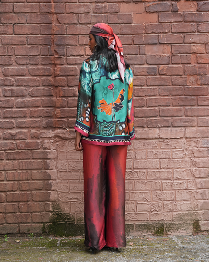 A model stands with their back to the camera against a rustic red-brown brick wall, wearing a vibrant turquoise and orange patterned blouse with red and black wide-leg pants and a matching red headscarf.