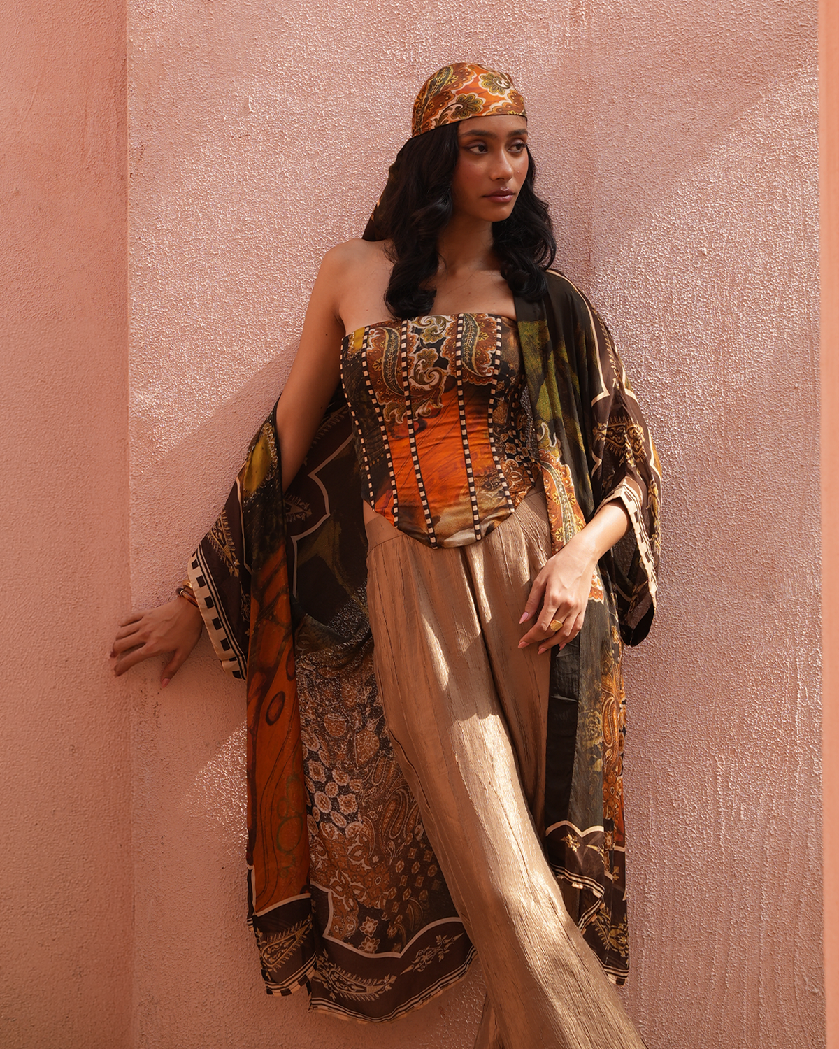 A woman stands against a textured peach-pink wall wearing a strapless bohemian-style top with an orange corset panel, wide beige trousers, a multicolored patterned shawl draped over her shoulders, and a matching patterned headscarf.