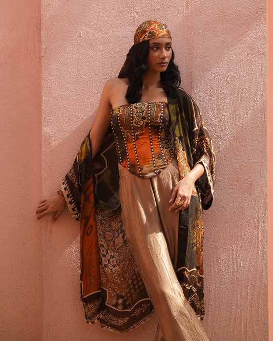 A woman stands against a textured peach-pink wall wearing a strapless bohemian-style top with an orange corset panel, wide beige trousers, a multicolored patterned shawl draped over her shoulders, and a matching patterned headscarf.