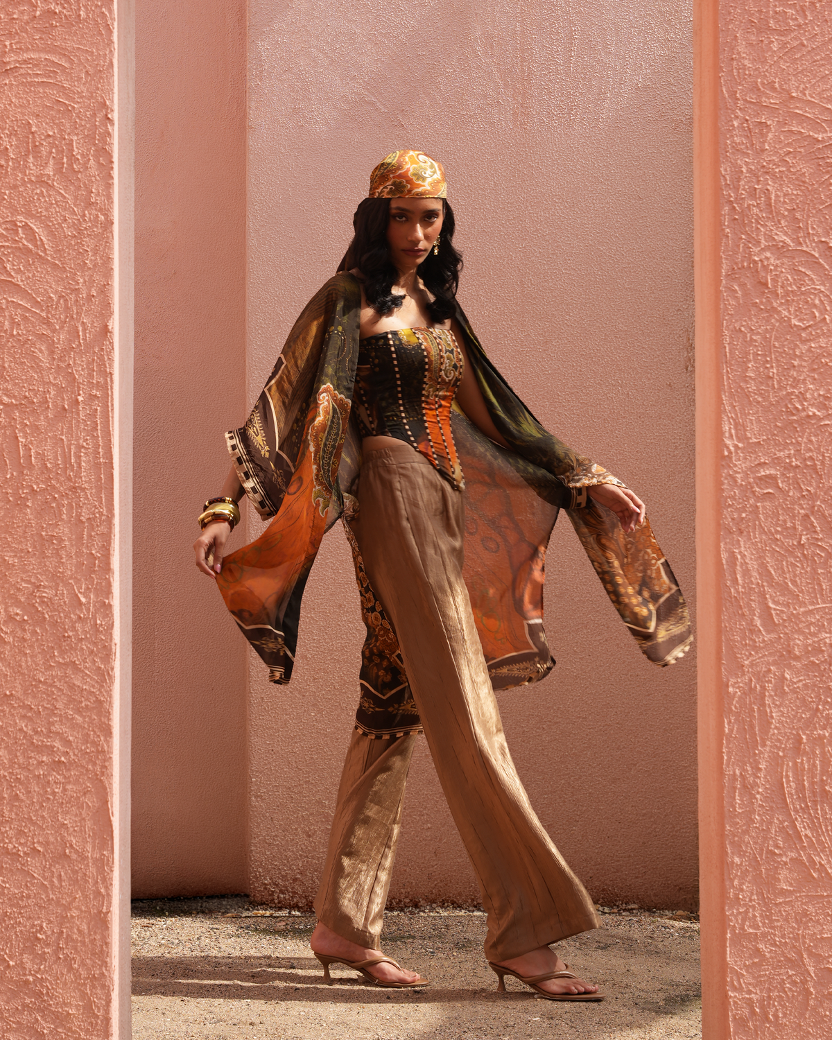 A woman models a traditional Moroccan-style outfit consisting of a black embroidered corset top, wide-leg beige trousers, a sheer patterned orange and black shawl, and a matching patterned headscarf, framed by a pink textured doorway.