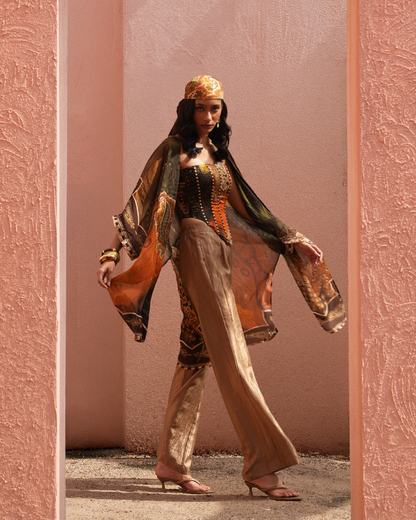 A woman models a traditional Moroccan-style outfit consisting of a black embroidered corset top, wide-leg beige trousers, a sheer patterned orange and black shawl, and a matching patterned headscarf, framed by a pink textured doorway.