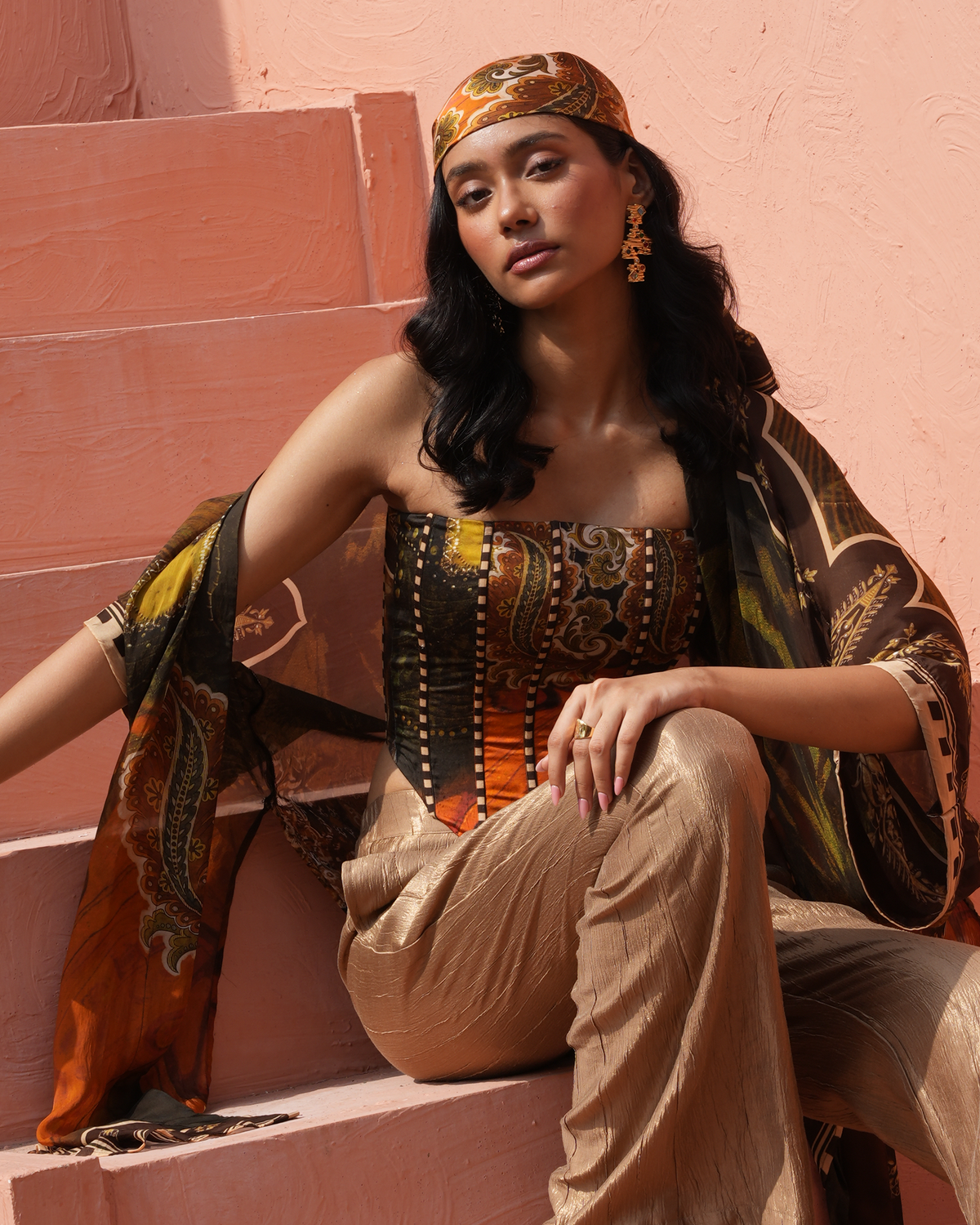 A woman sits on pink outdoor steps wearing a coordinated printed outfit with a patterned bandeau-style top, loose beige pants, a matching headscarf, and a draped scarf. The styling includes visible jewelry, and the background is a pink stucco wall.