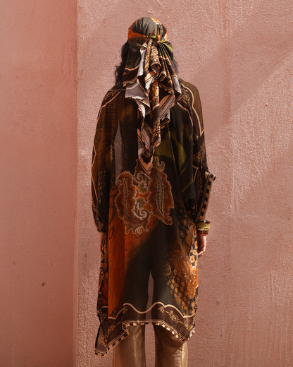 A person viewed from behind wearing a loose, long printed tunic or kaftan with a matching headscarf, standing against a textured pink wall. The garment has an ornate multicolor paisley and floral print in black, brown, orange, beige, and muted green tones.