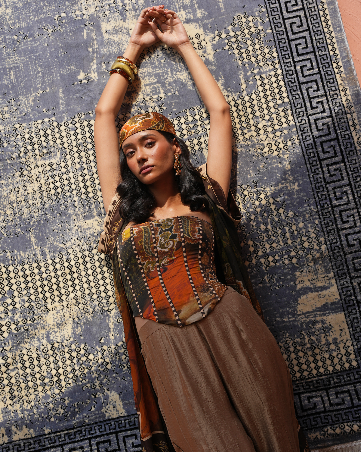 A woman wearing a bohemian-style outfit with a patterned corset top, brown skirt, and headband, posing with her arms raised against a blue and beige patterned rug.