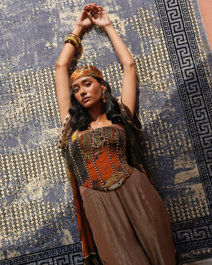 A woman wearing a bohemian-style outfit with a patterned corset top, brown skirt, and headband, posing with her arms raised against a blue and beige patterned rug.
