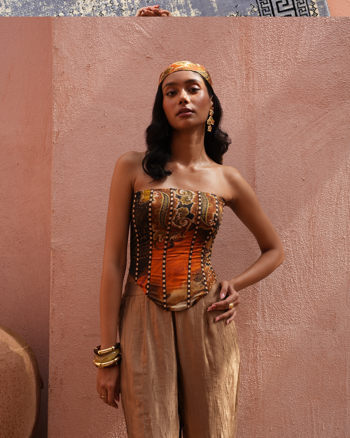 A woman models a Moroccan-style strapless corset top with intricate gold, brown, and orange patterns, paired with matching beige trousers and a gold headband, set against a textured terracotta wall.