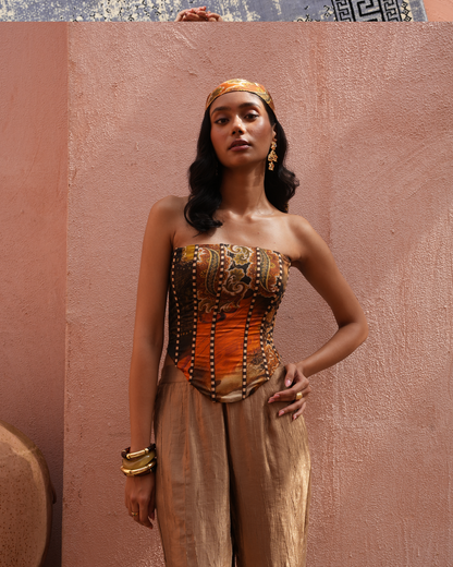 A woman models a Moroccan-style strapless corset top with intricate gold, brown, and orange patterns, paired with matching beige trousers and a gold headband, set against a textured terracotta wall.