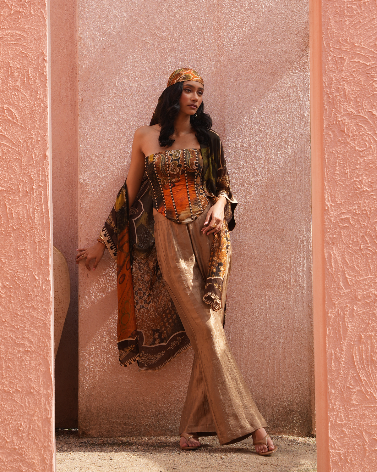 A woman stands in a doorway wearing a bohemian-style outfit with a patterned orange and brown corset top, matching draped jacket, beige wide-leg pants, and a headscarf against a pink textured wall.