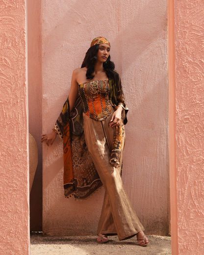 A woman stands in a doorway wearing a bohemian-style outfit with a patterned orange and brown corset top, matching draped jacket, beige wide-leg pants, and a headscarf against a pink textured wall.