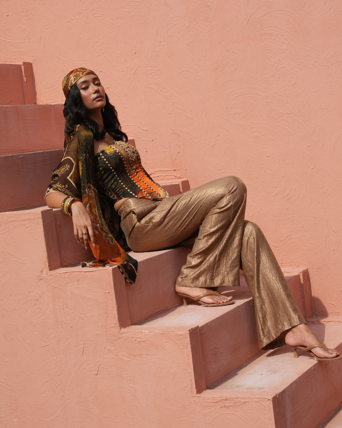 A woman models a bohemian-style outfit consisting of metallic bronze pants, a black and orange patterned top, and a matching patterned headscarf while sitting on terracotta-colored steps against a textured pink wall.