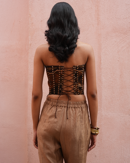 A woman viewed from behind wearing a brown strapless lace-up corset top and tan drawstring pants, standing against a textured pink wall.