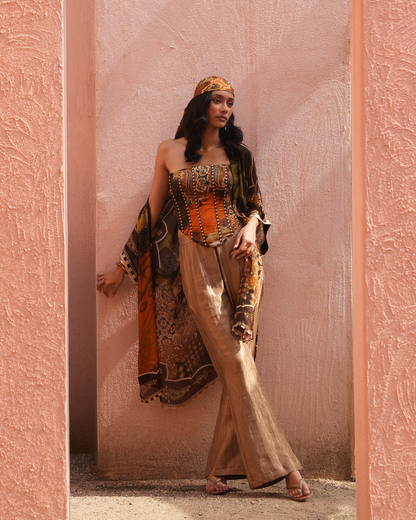 A woman models a bohemian-style outfit consisting of a metallic gold corset top, wide-leg metallic trousers, a patterned orange and black shawl, and a matching headscarf, posing against a textured pink stucco wall.