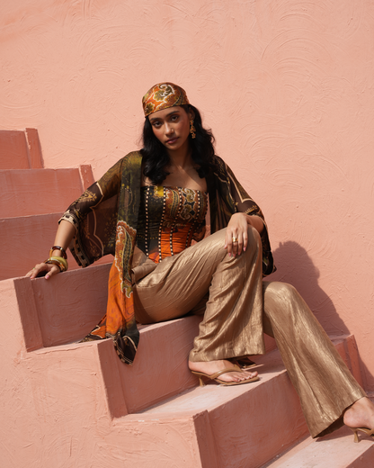 A woman sits on terracotta-colored steps against a matching textured wall, wearing a metallic gold and orange outfit with a patterned headscarf and draped scarf.