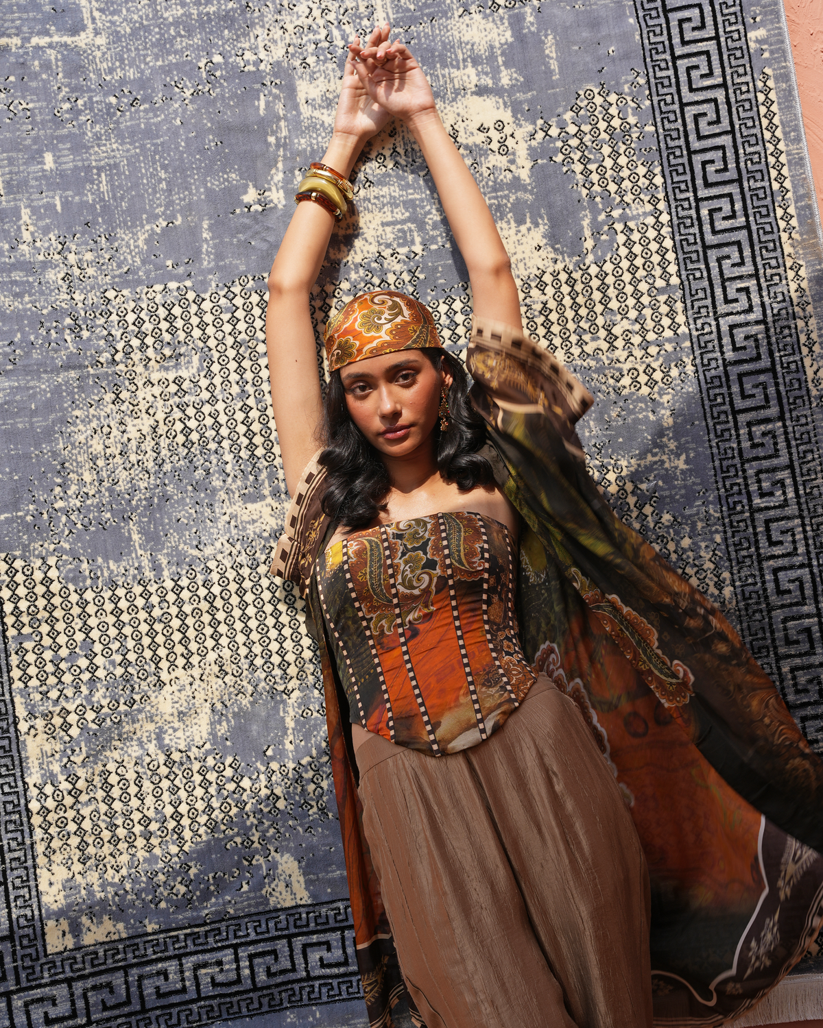 A woman posing in an ornate, bohemian-style costume featuring a brown and orange embroidered corset top, a brown skirt, a patterned headband, and a draped scarf, set against a patterned blue and gold rug.