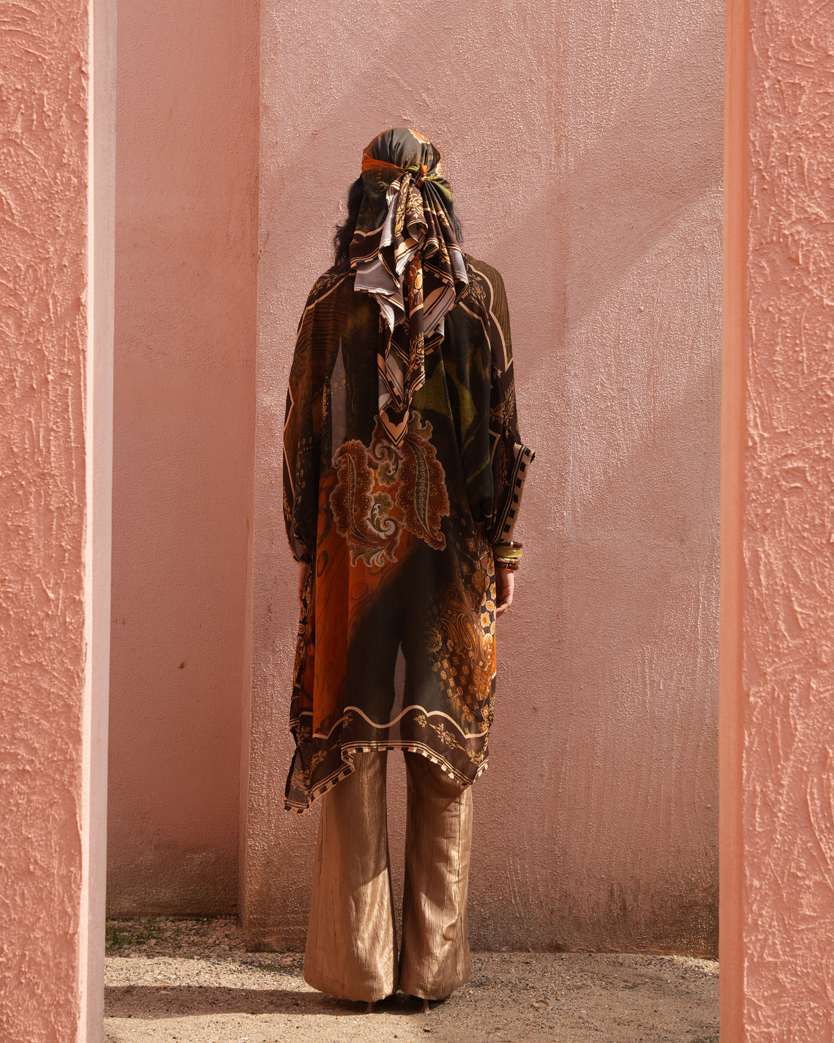 A model viewed from behind wearing a long-sleeved, loose-fitting printed tunic in earthy tones of brown, orange, and green, paired with metallic gold wide-leg trousers and a patterned headscarf, standing in a doorway against a textured pink wall.