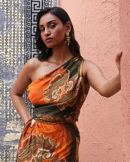 A woman wearing a one-shoulder dress with an orange, brown, and green paisley print, posing against a textured peach-pink wall next to a patterned blue and gray tile panel.