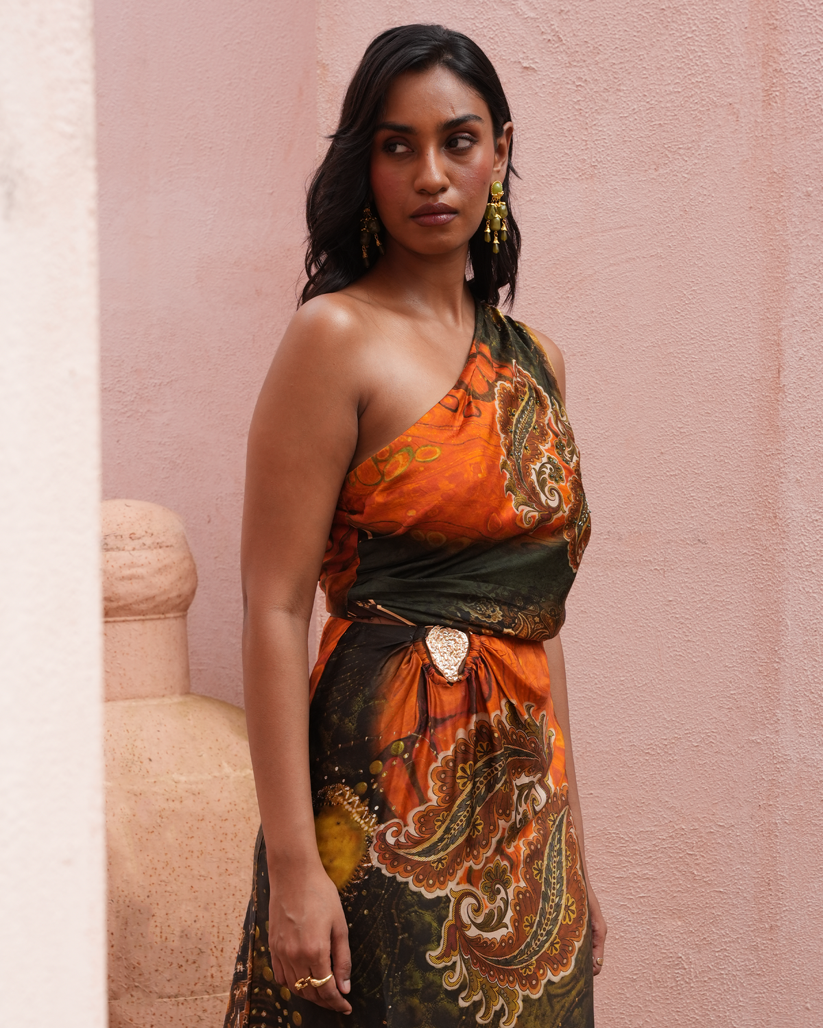 A woman wearing a one-shoulder printed dress with orange, black, and gold paisley patterns, standing against a textured pink wall.