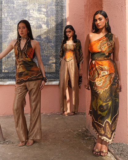 Three women model matching printed outfits against a pink stucco wall and blue patterned tapestry. The garments feature a rich mix of orange, brown, black, and gold paisley and floral motifs.