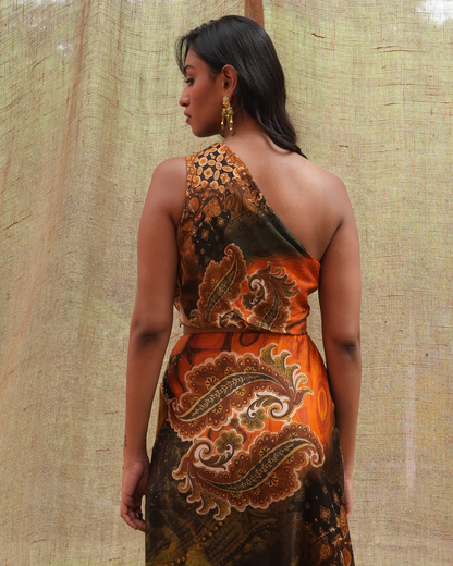 A woman shown from the back wearing a one-shoulder dress with a bold paisley print in orange, brown, and black, posed against a textured beige fabric backdrop.