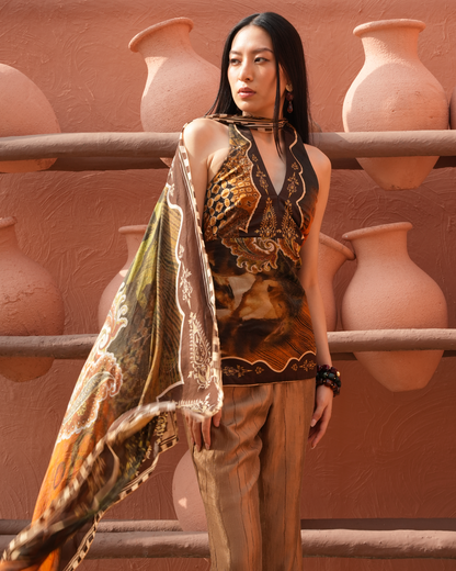 A woman stands in front of a terracotta wall with large clay pots, wearing a sleeveless halter top and matching pants in a brown and gold paisley print, with a patterned scarf draped over one shoulder.