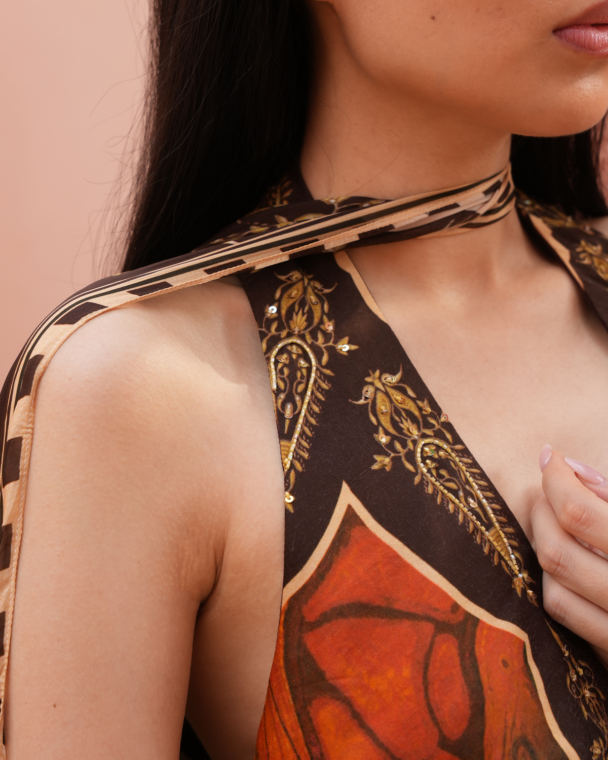 Close-up of a model wearing a halter-style top or dress made from a patterned scarf, featuring a brown paisley print with gold detailing and a rust-orange abstract section, against a soft peach background.