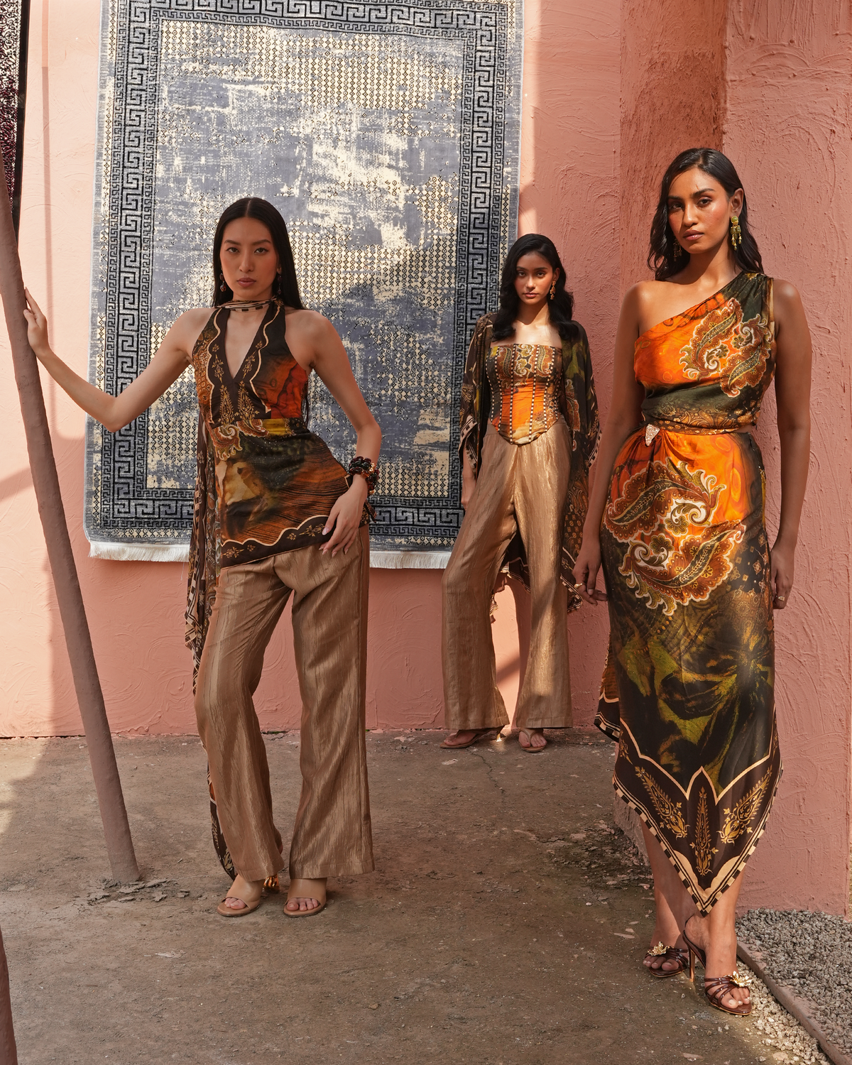 Three models wearing coordinated earth-toned printed clothing stand against a pink stucco wall with a large blue patterned textile hanging behind them. The outfits feature draped, painterly prints in orange, brown, black, and beige.