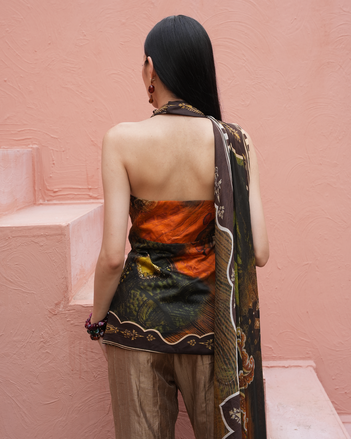 A woman is shown from behind wearing a strapless halter-style top with a colorful landscape print in orange, green, and brown, paired with beige trousers and a matching printed scarf, set against a textured pink wall.