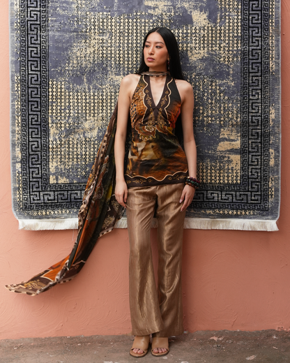 A woman stands against a peach-colored wall wearing a sleeveless halter top and wide-leg metallic pants, with a matching patterned scarf draped over her shoulder. A large dark patterned rug hangs on the wall behind her.