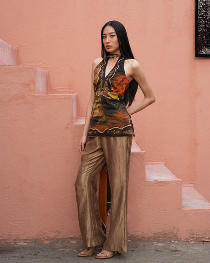 A woman stands outdoors against a textured peach-pink wall with steps, wearing a sleeveless halter-style top with a dark abstract print in orange, brown, and black, paired with wide-leg metallic bronze pants.