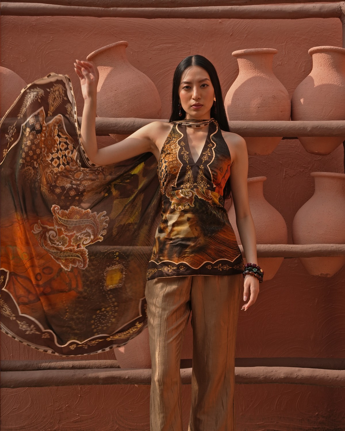 A woman stands in front of a terracotta wall with large clay pots, wearing a sleeveless patterned halter-style top and matching brown pants, while holding a sheer patterned scarf.
