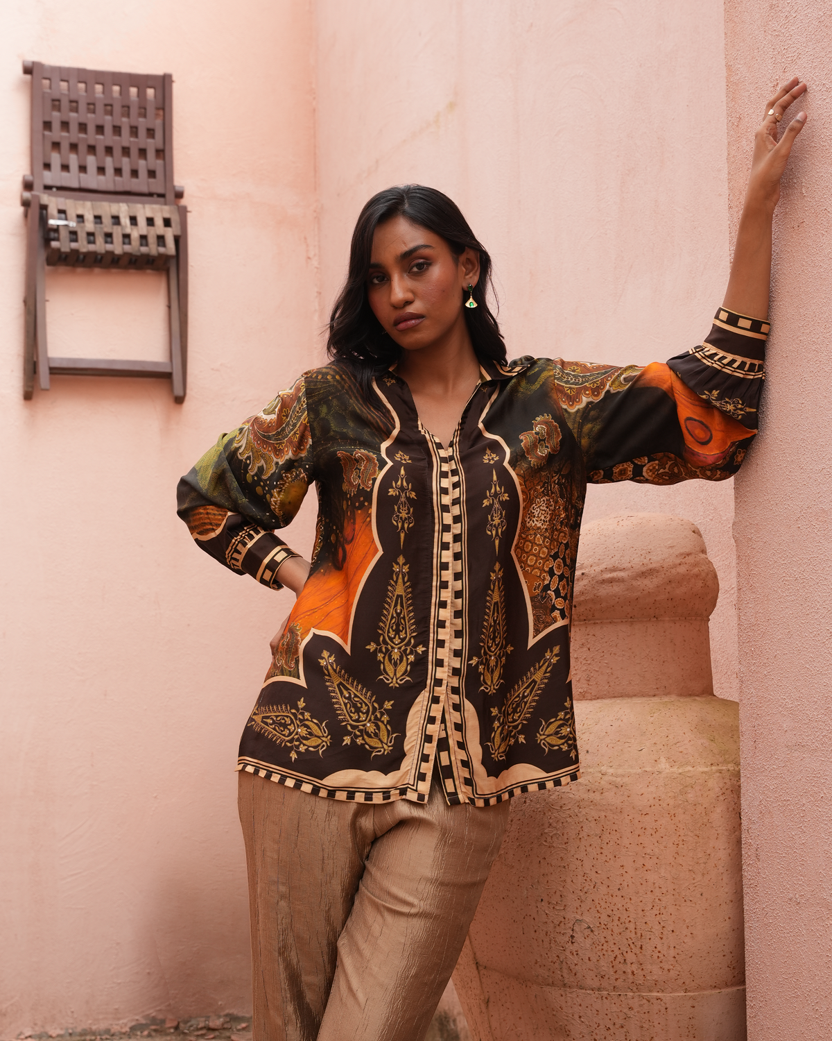A woman models a long-sleeve button-up shirt with an intricate black, gold, and orange paisley pattern, paired with beige trousers, posing against a textured pink wall.