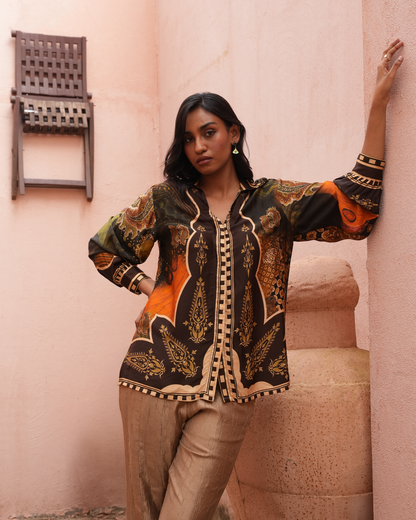 A woman models a long-sleeve button-up shirt with an intricate black, gold, and orange paisley pattern, paired with beige trousers, posing against a textured pink wall.