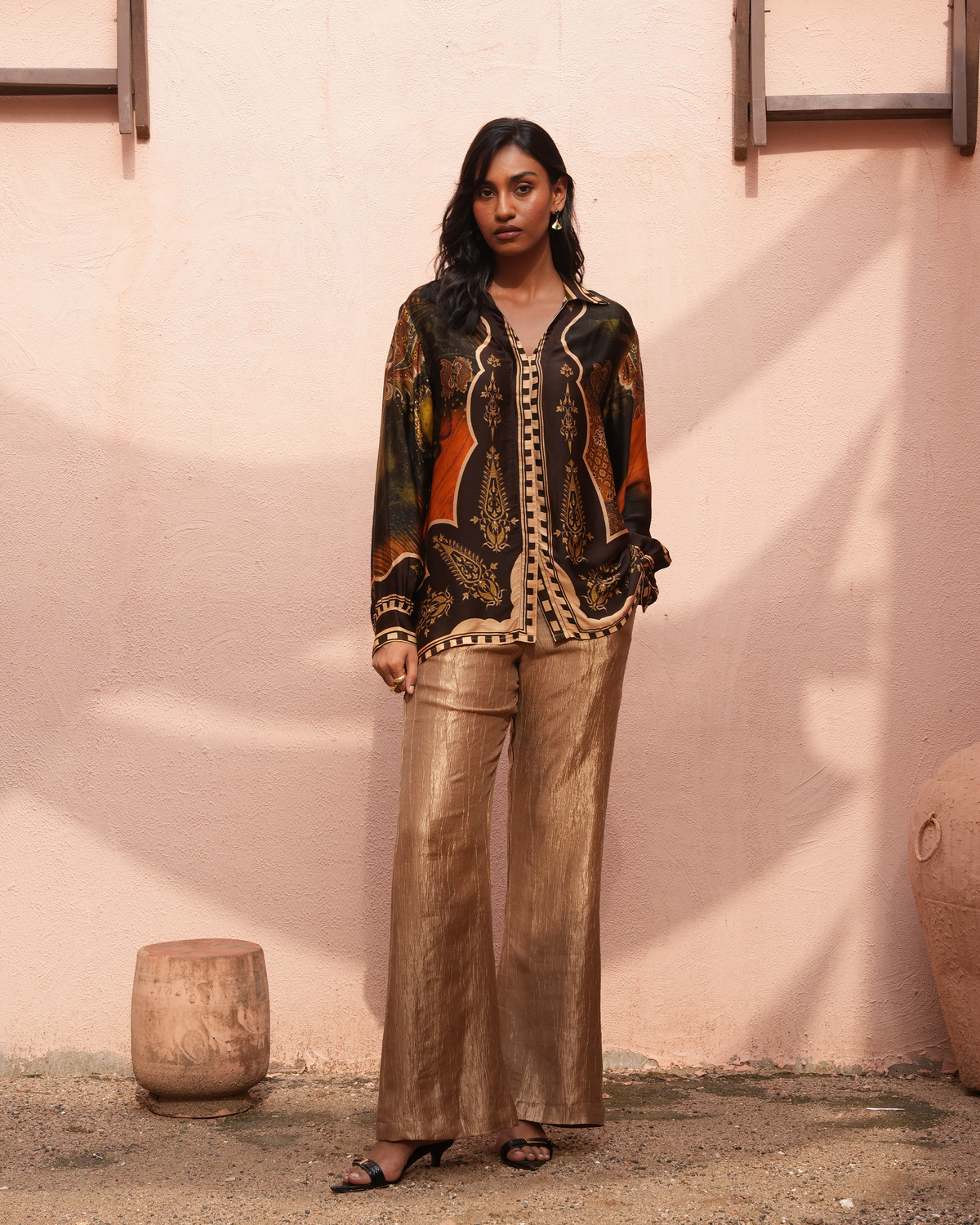 A woman stands outdoors against a textured pink wall wearing a black and orange patterned button-up shirt with metallic gold wide-leg pants and black sandals.