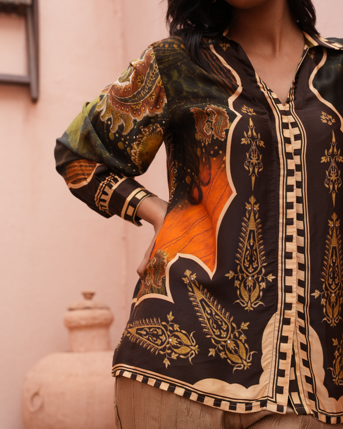 A woman wearing a long-sleeve button-up shirt with an intricate paisley and floral print in black, orange, green, and beige, posed against a soft pink wall.
