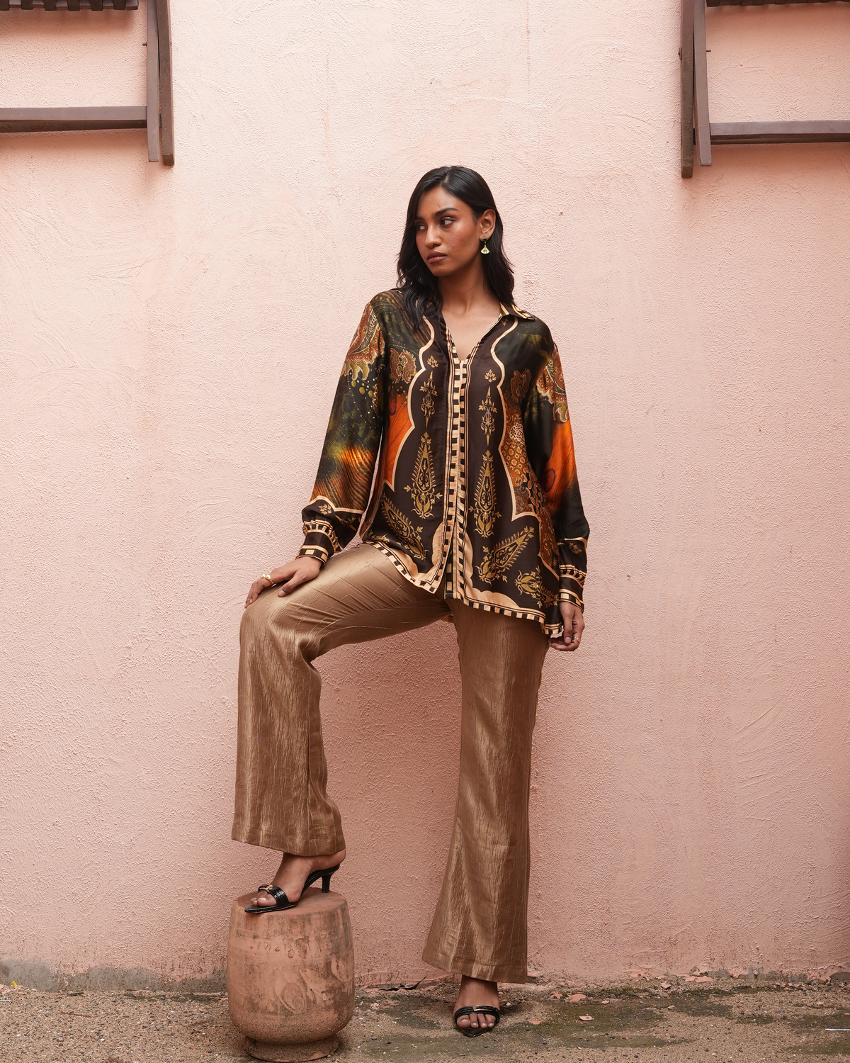 A woman models a bohemian-style two-piece outfit consisting of a long-sleeve button-up shirt with a multicolor paisley and tribal print, and matching metallic gold flared trousers, posing against a textured pink wall.