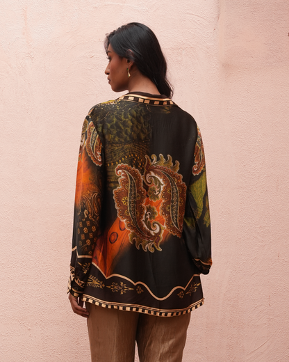 A woman shown from the back wearing a long-sleeve black silk-like blouse with a large orange, gold, and green paisley print, standing against a textured light pink wall.