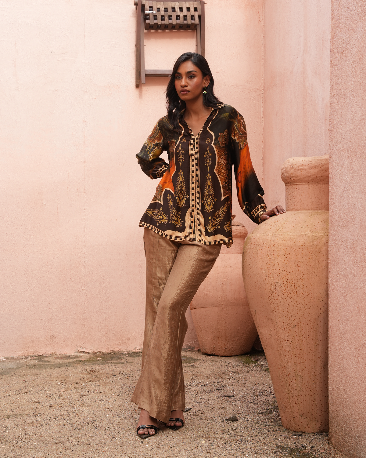 A woman models a two-piece traditional outfit consisting of a black and orange printed long-sleeve top with gold detailing and matching beige flared trousers, standing in front of a pink stucco wall with large terracotta pots.