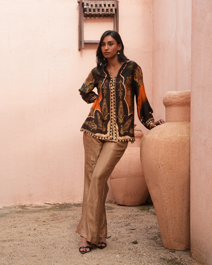 A woman models a two-piece traditional outfit consisting of a black and orange printed long-sleeve top with gold detailing and matching beige flared trousers, standing in front of a pink stucco wall with large terracotta pots.