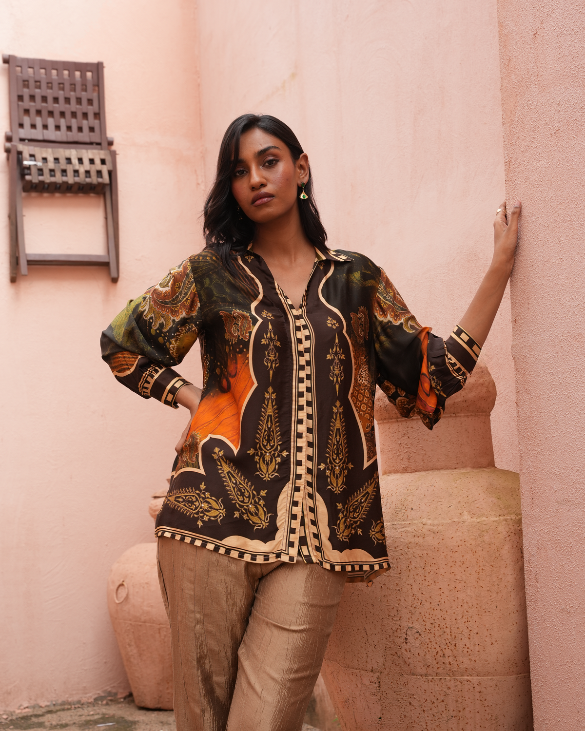 A woman models a long-sleeve button-up shirt with a black base and intricate gold, beige, and orange paisley patterns, paired with beige trousers, set against a warm pink stucco wall.