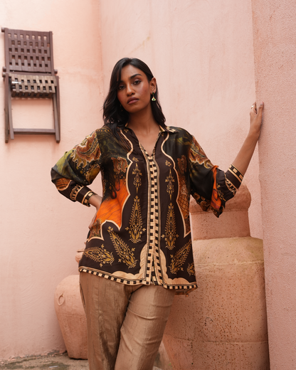 A woman models a long-sleeve button-up shirt with a black base and intricate gold, beige, and orange paisley patterns, paired with beige trousers, set against a warm pink stucco wall.